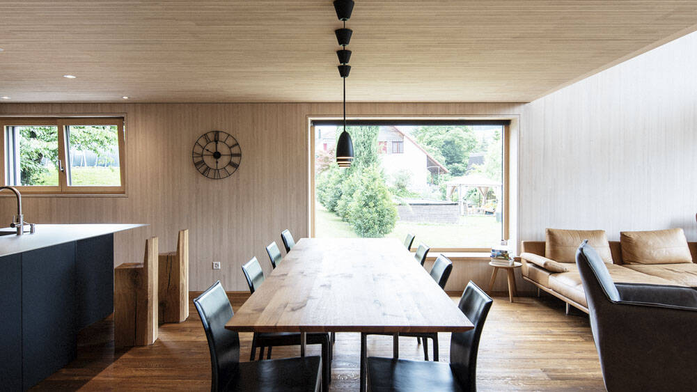 Die lange Essentafel steht direkt gegenüber einer schaufensterartigen, quadratischen Fensterfront und gewährt den Sitzenden so einen Blick in die Natur. 