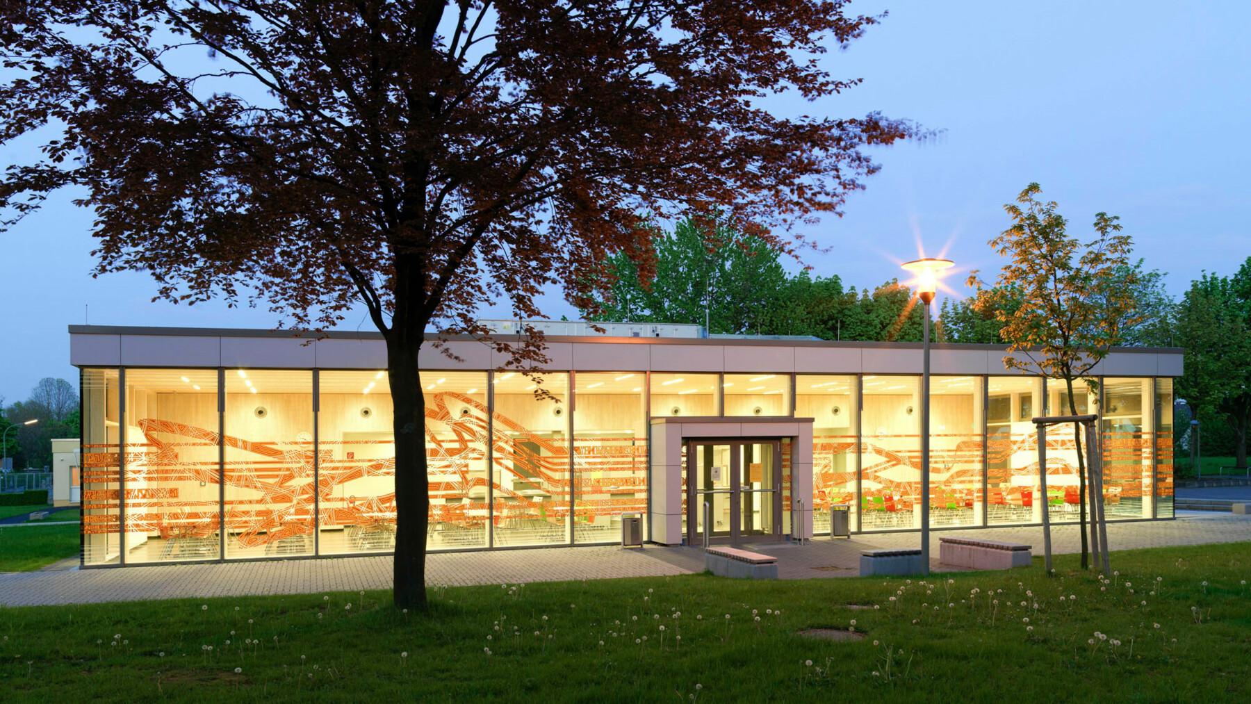 Die Mensa des Gymnasiums in Paderborn wird zur Westseite hin durch eine große Glasfassade mit Bemalungen in sanfter Erdfarbe begrenzt.