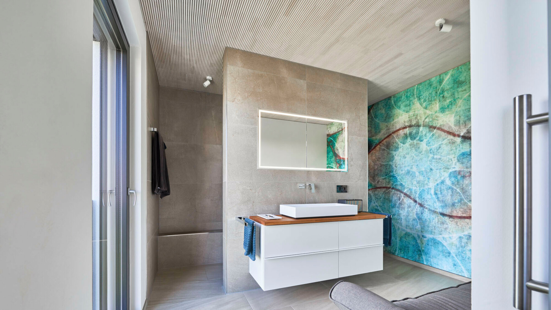 Dieses Badezimmer bekommt einen besonderen Flair durch die kunstvoll bedruckte Wand in Blau-und Grüntönen, sowie die feine Deckenprofilierung, die eine dynamische Atmosphäre kreiert. 