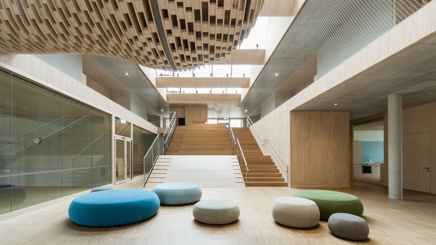 Die breite tribünenartige Treppenanlage bildet die Verbindung zwischen Foyer und OG. Farbige Sitzpolster lockern den von Holz, Glas und Metall dominierten Raum auf.