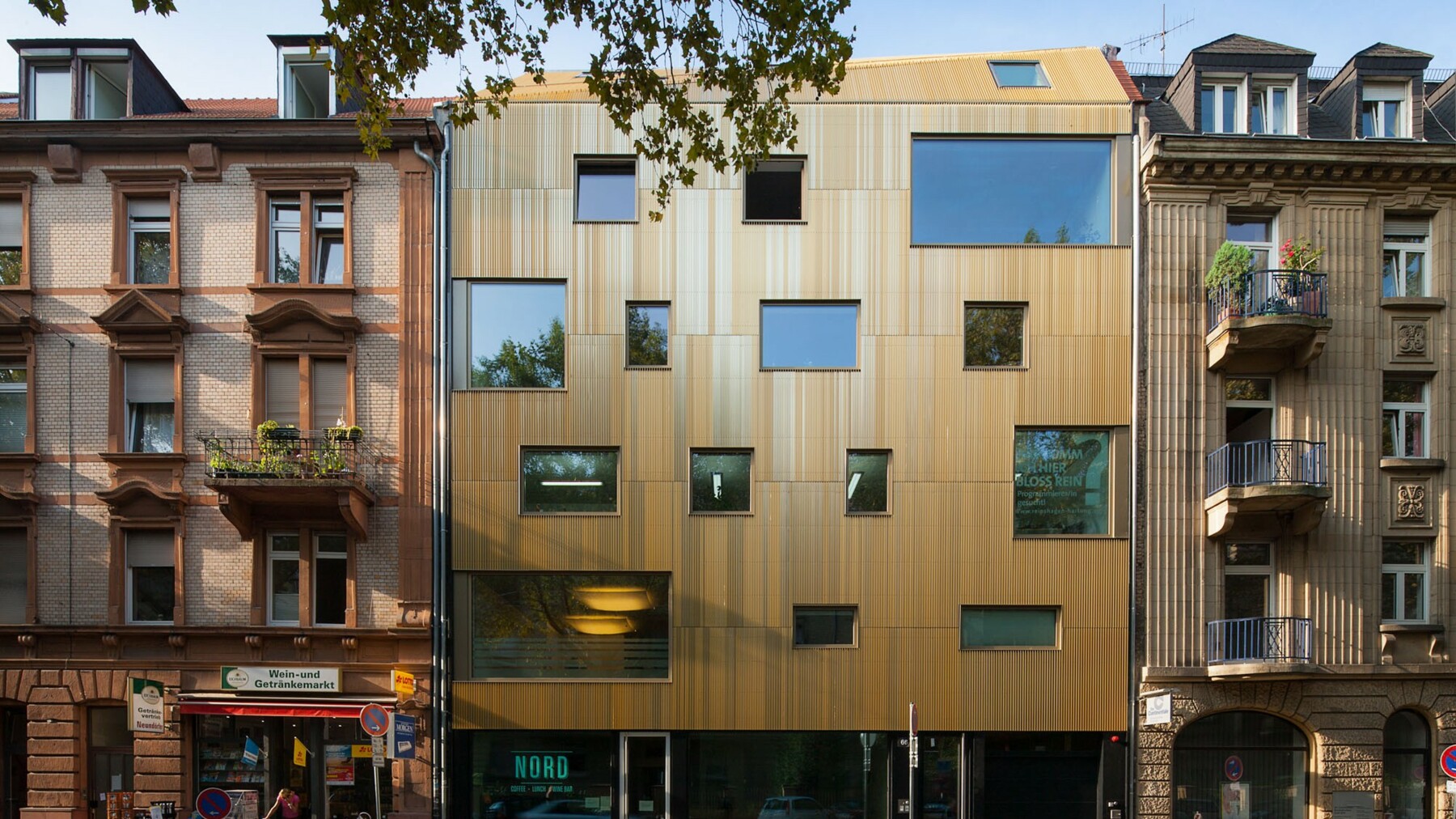 Die Außenansicht dieses energieeffizienten Neubaus ist zwischen den denkmalgeschützten Altbauten mit den unterschiedlich großen Fenstern und der Fassade in Gold-und Hellbraun-Tönen ein echter Blickfang. 